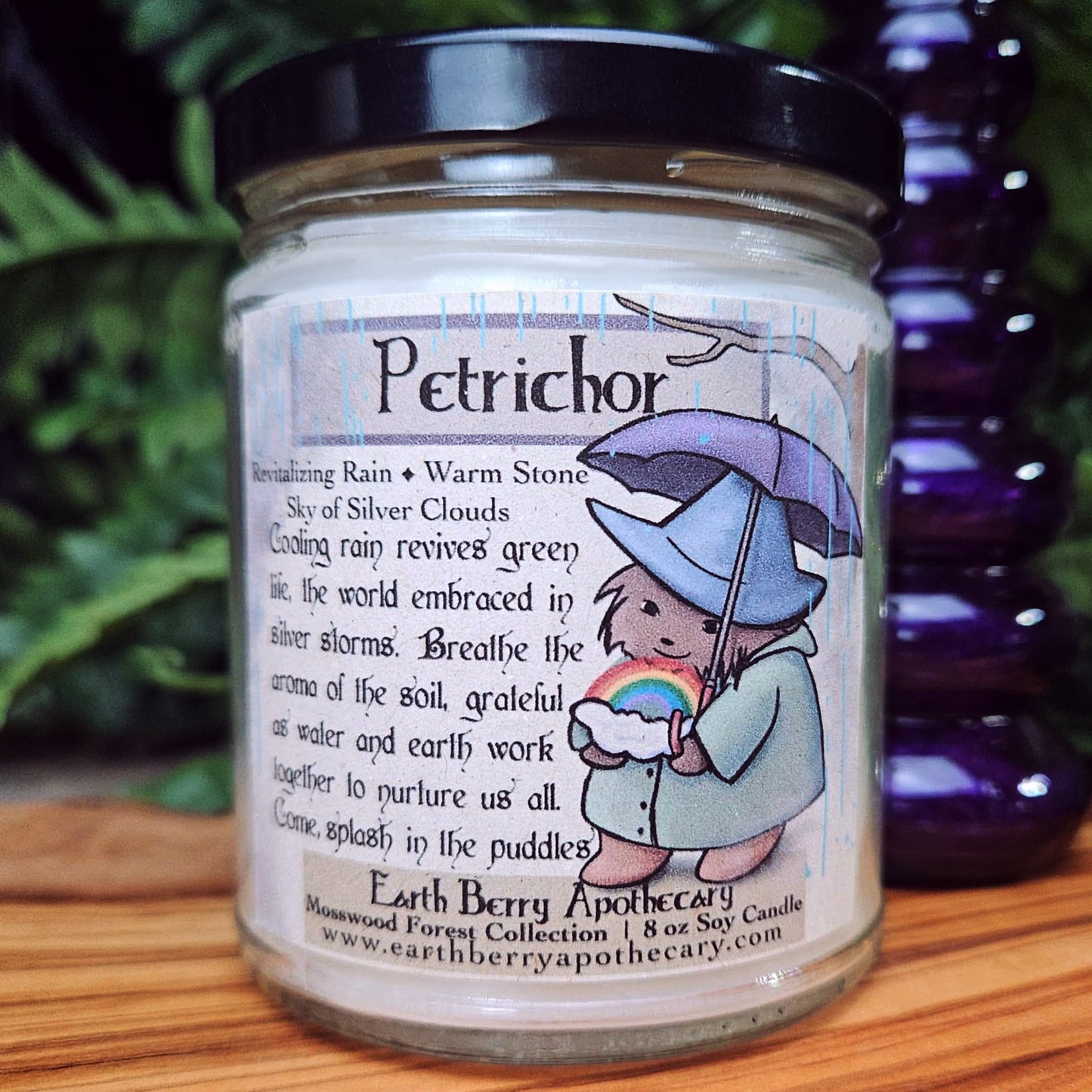 Candle jar labeled 'Petrichor' with a hedgehog holding an umbrella and rainbow design, set against a natural background.