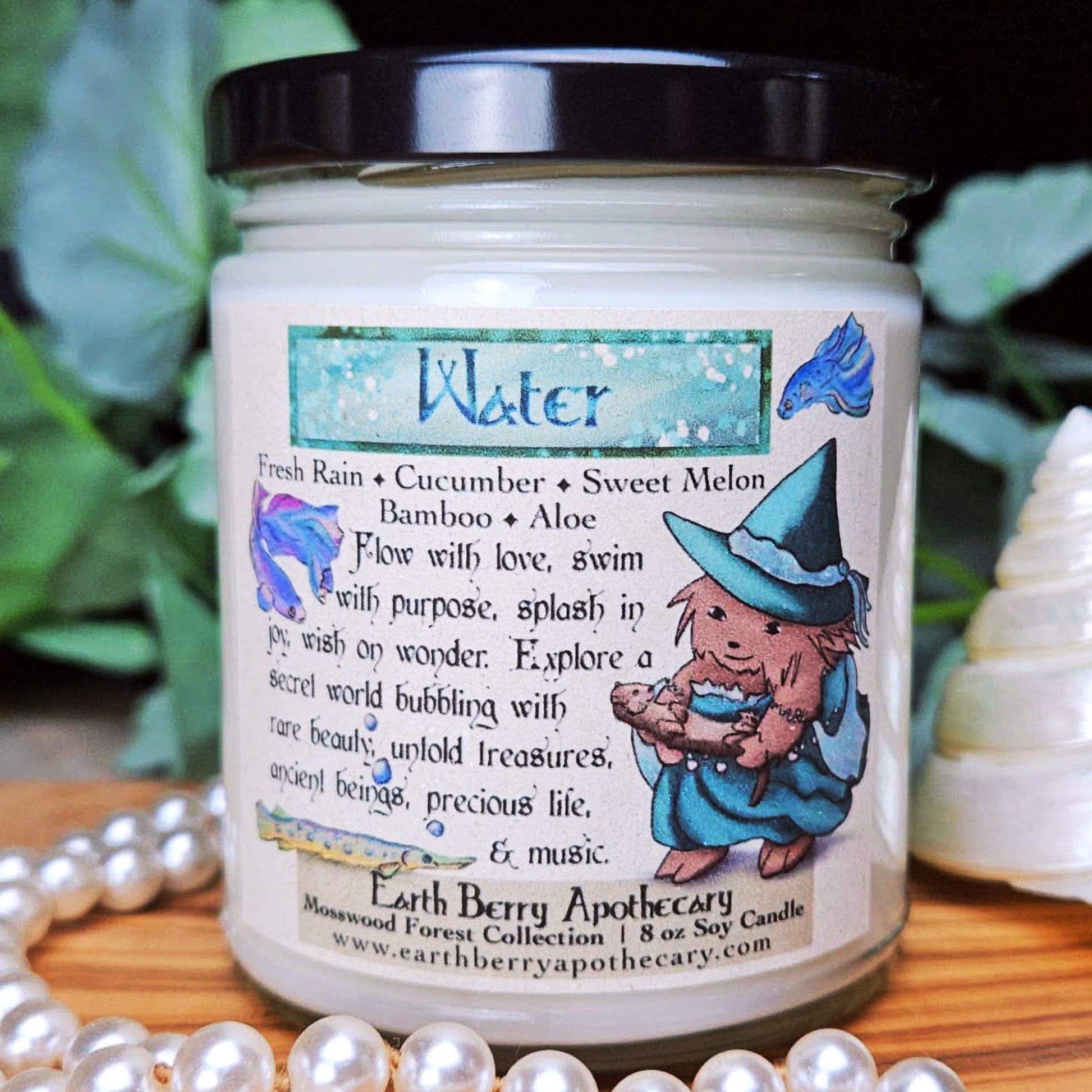 Candle jar with 'Water' theme and a hedgehog witch illustration with beta fish, an otter, and a gar. The candle sits on a wooden surface with pearls, shells, and greenery.