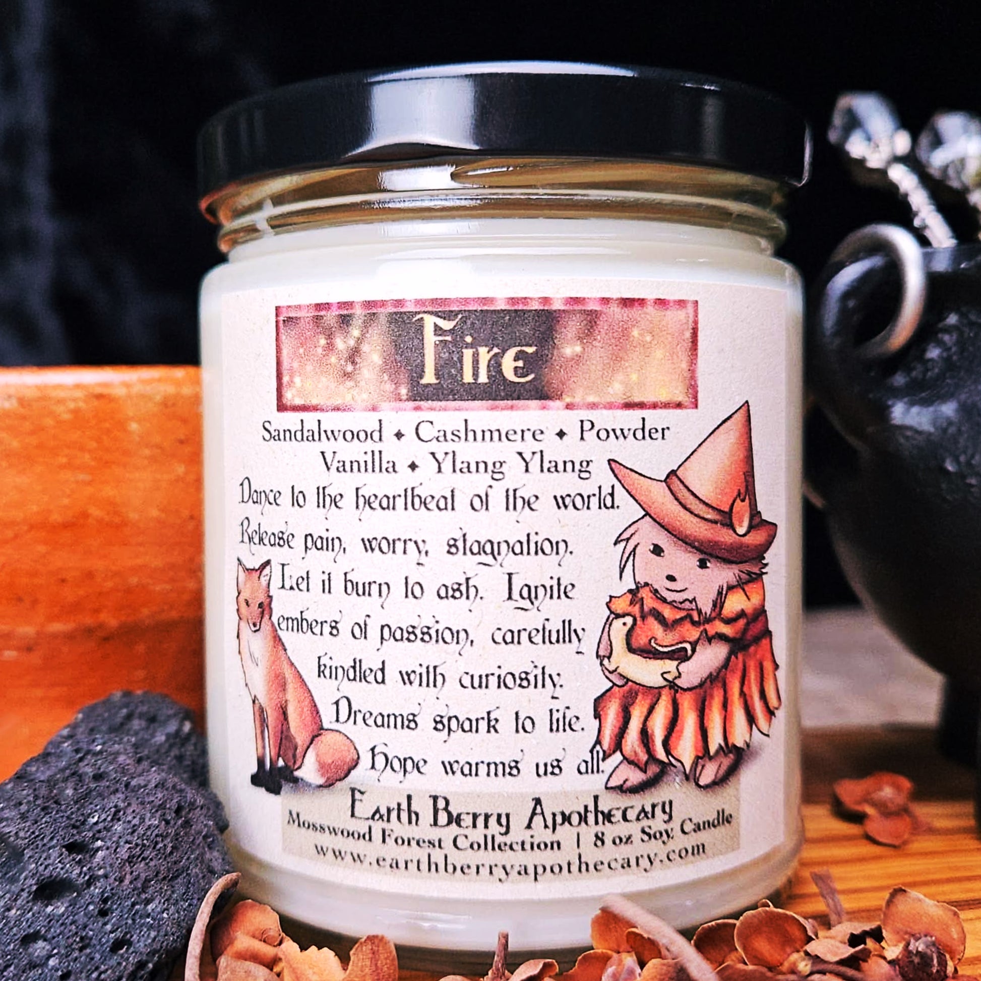 Candle jar with 'Fire' scent and label featuring a hedgehog witch and fox illustration, on a dark background. The scent is vanilla cashmere and sandalwood.