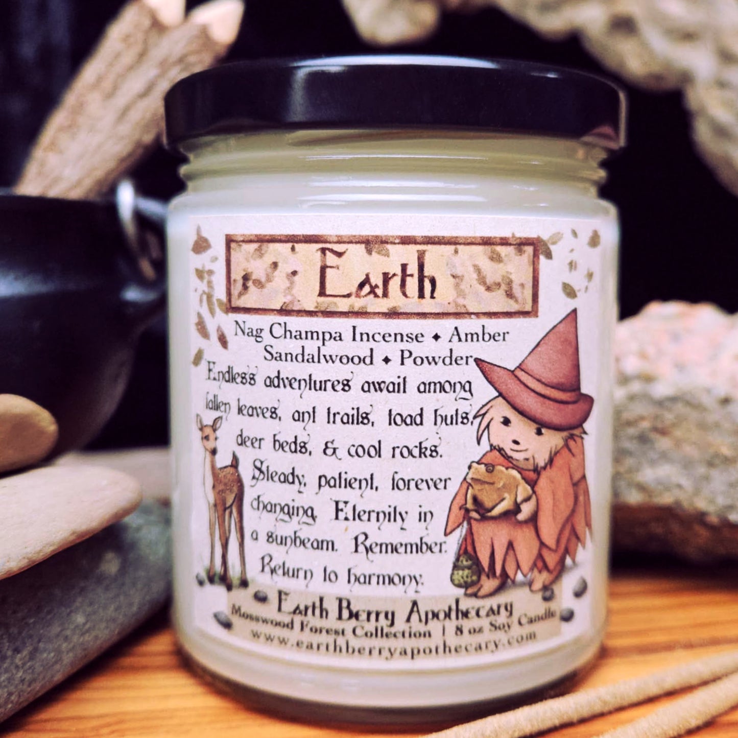 Jar of Earth Berry Apothecary candle called Earth with a hedgehog witch, toad, and deer illustration on a wooden surface surrounded by stones and sticks.