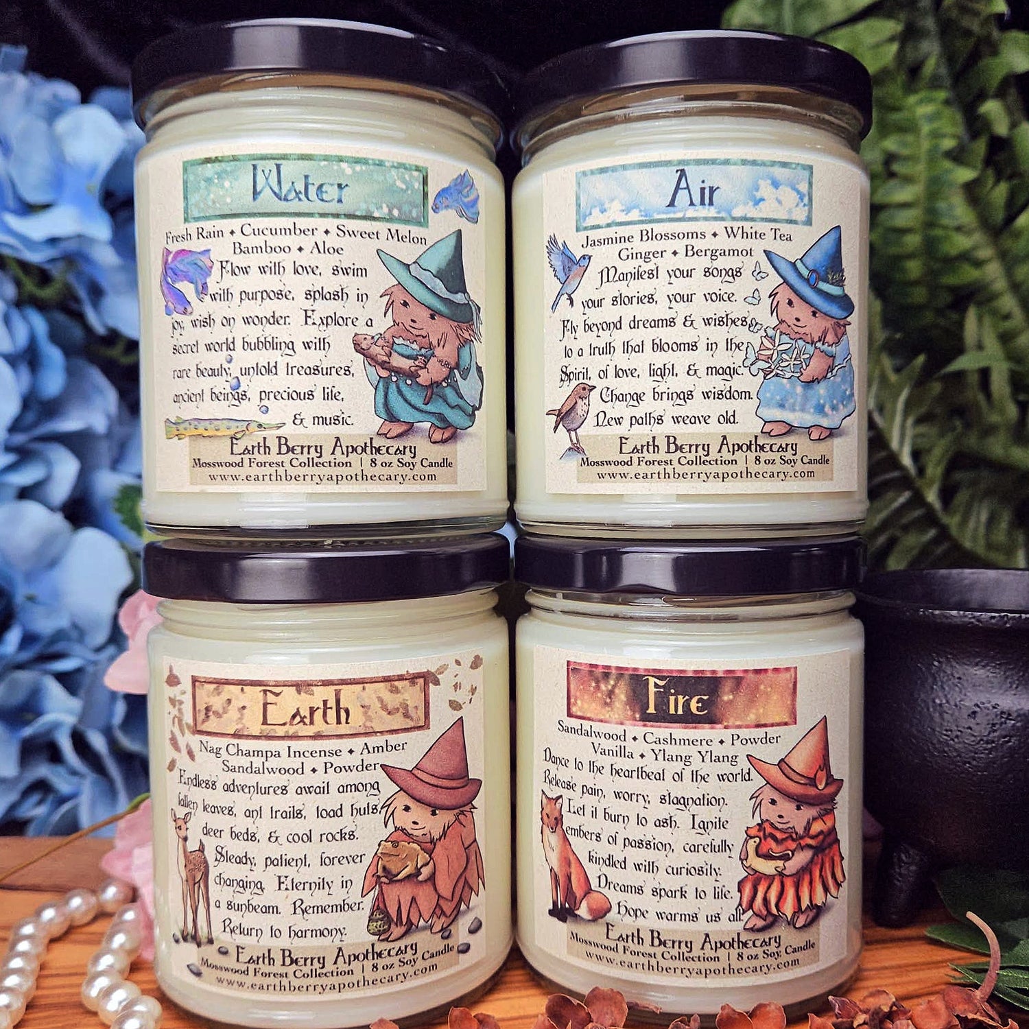 Four candles with labels representing Water, Air, Earth, and Fire, stacked on a wooden surface with a floral and fern background.