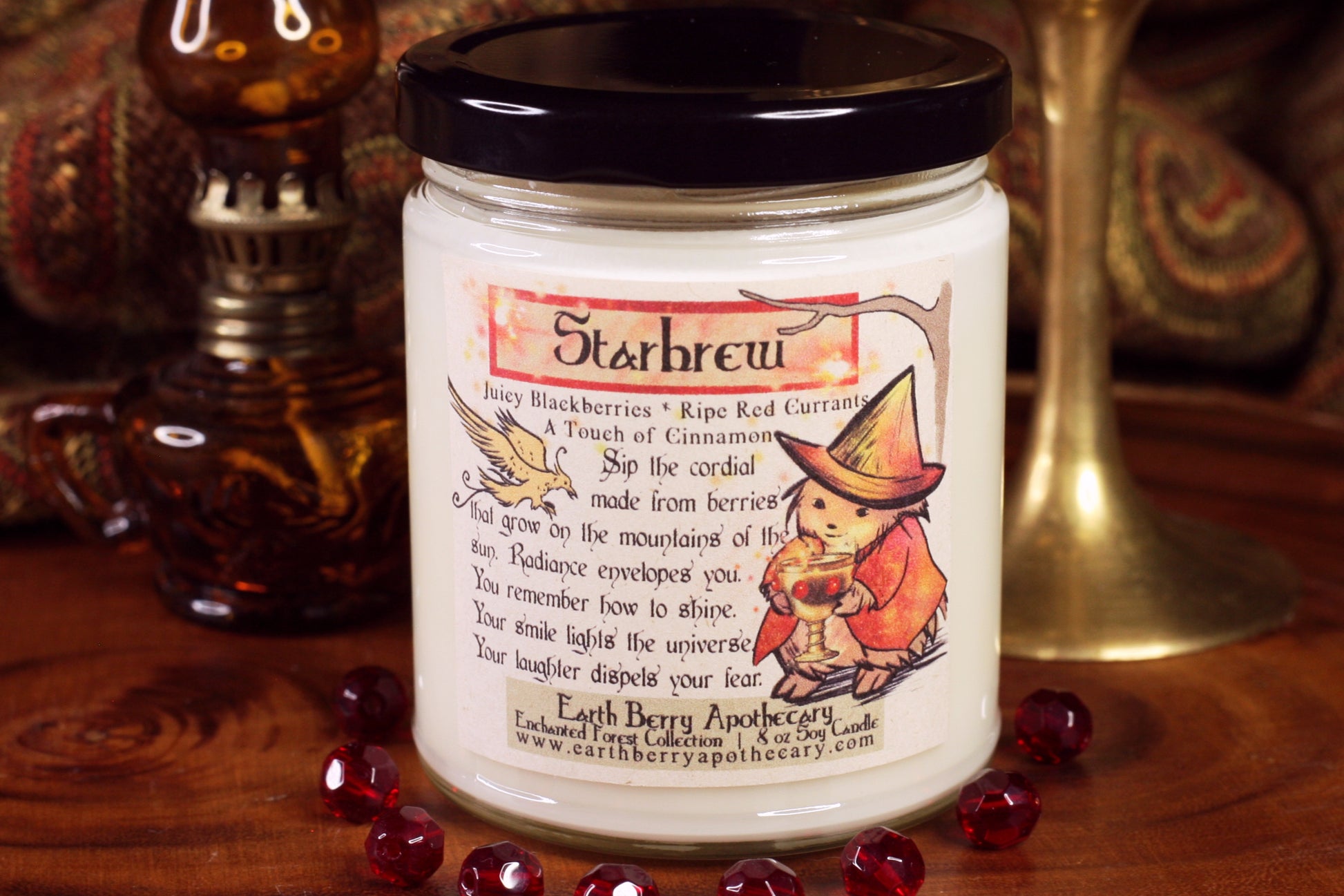 Starbrew soy candle with the hedge witch wearing a red jacket, holding a glowing golden goblet. A sun bird flies in from the side. The scent is red currants, oranges, citrus, and berries.