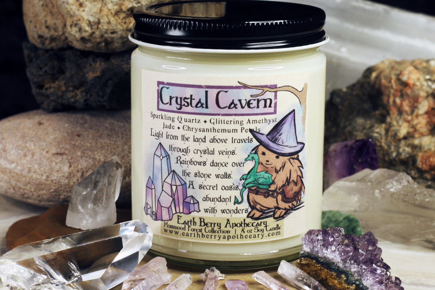 Crystal cavern soy candle with the hedge witch wearing a leather pouch, holding a baby dragon. Blue pink and purple crystals sparkle too. The scent is chrysanthemum, jade, quartz, amethyst, and cactus flower.