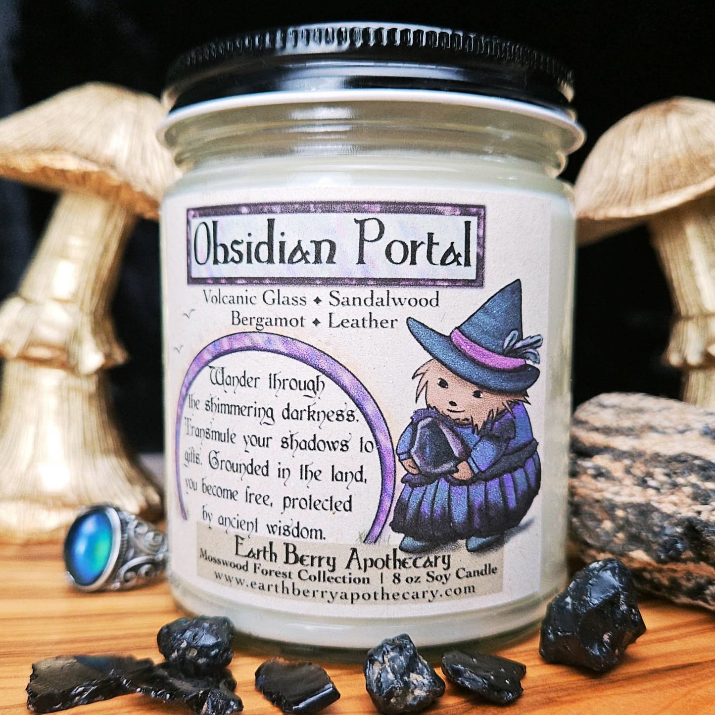 Candle jar labeled 'Obsidian Portal' featuring a cartoon hedgehog wearing a witch hat and clothes, with decorative stones on a wooden surface.