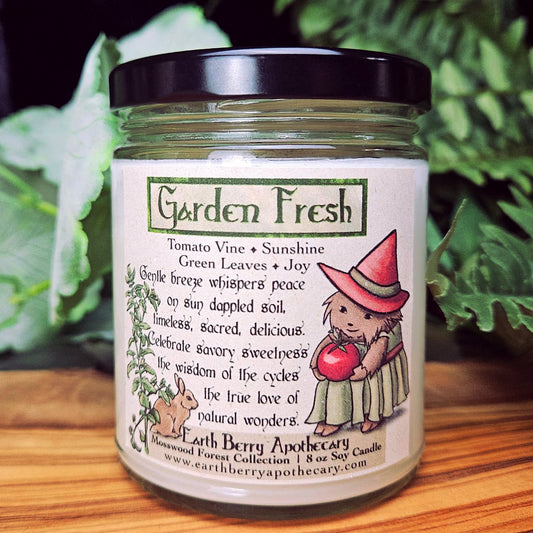 Candle jar labeled 'Garden Fresh' with a cartoon hedgehog wearing a witch hat and clothes on a wooden surface with greenery in the background. The scent is tomato vine.