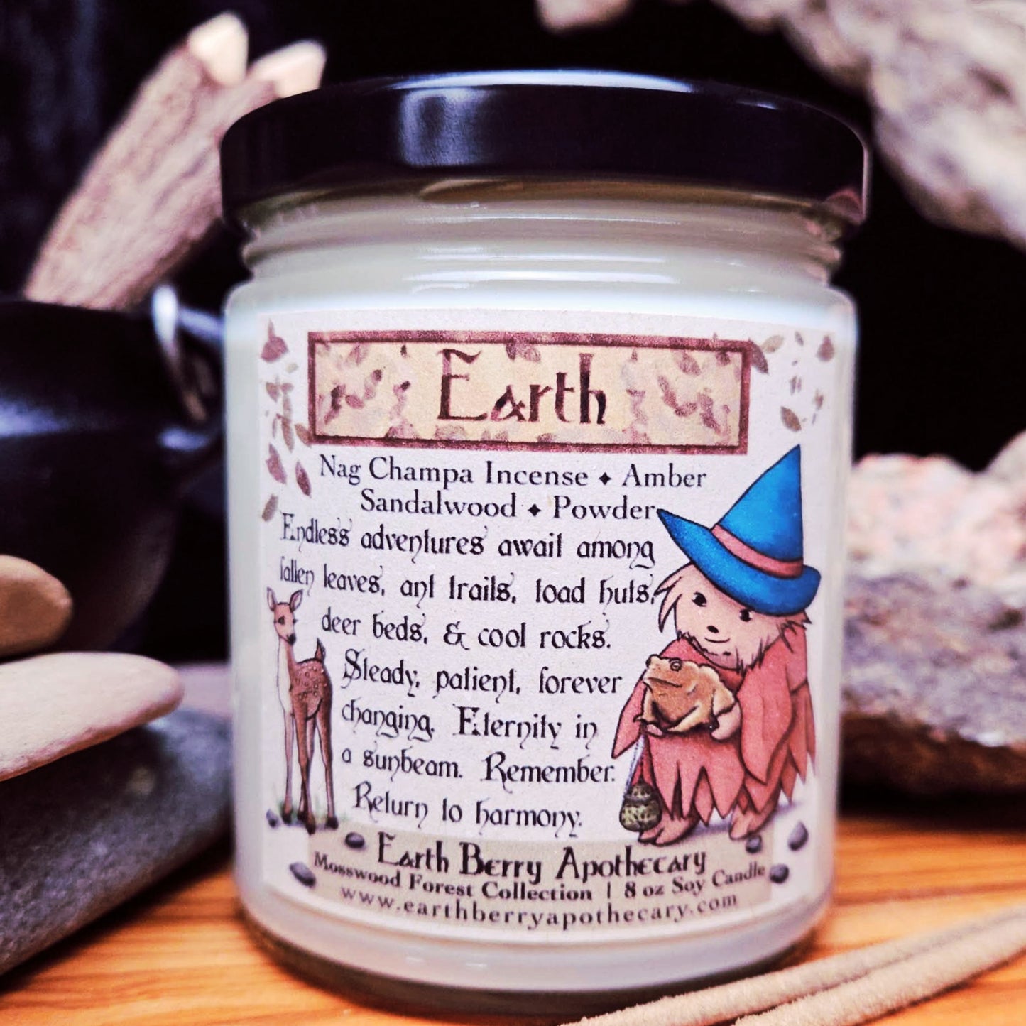 Candle jar labeled 'Earth' with a hedgehog witch and nature-themed design, surrounded by stones.